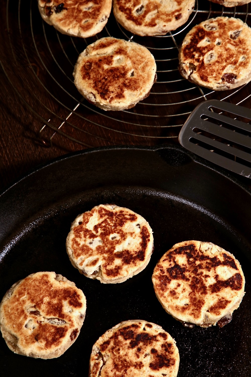 Four cooked vegan Welsh cakes in a black cast iron pan. Four more cakes are cooling on a wire rack in the background.