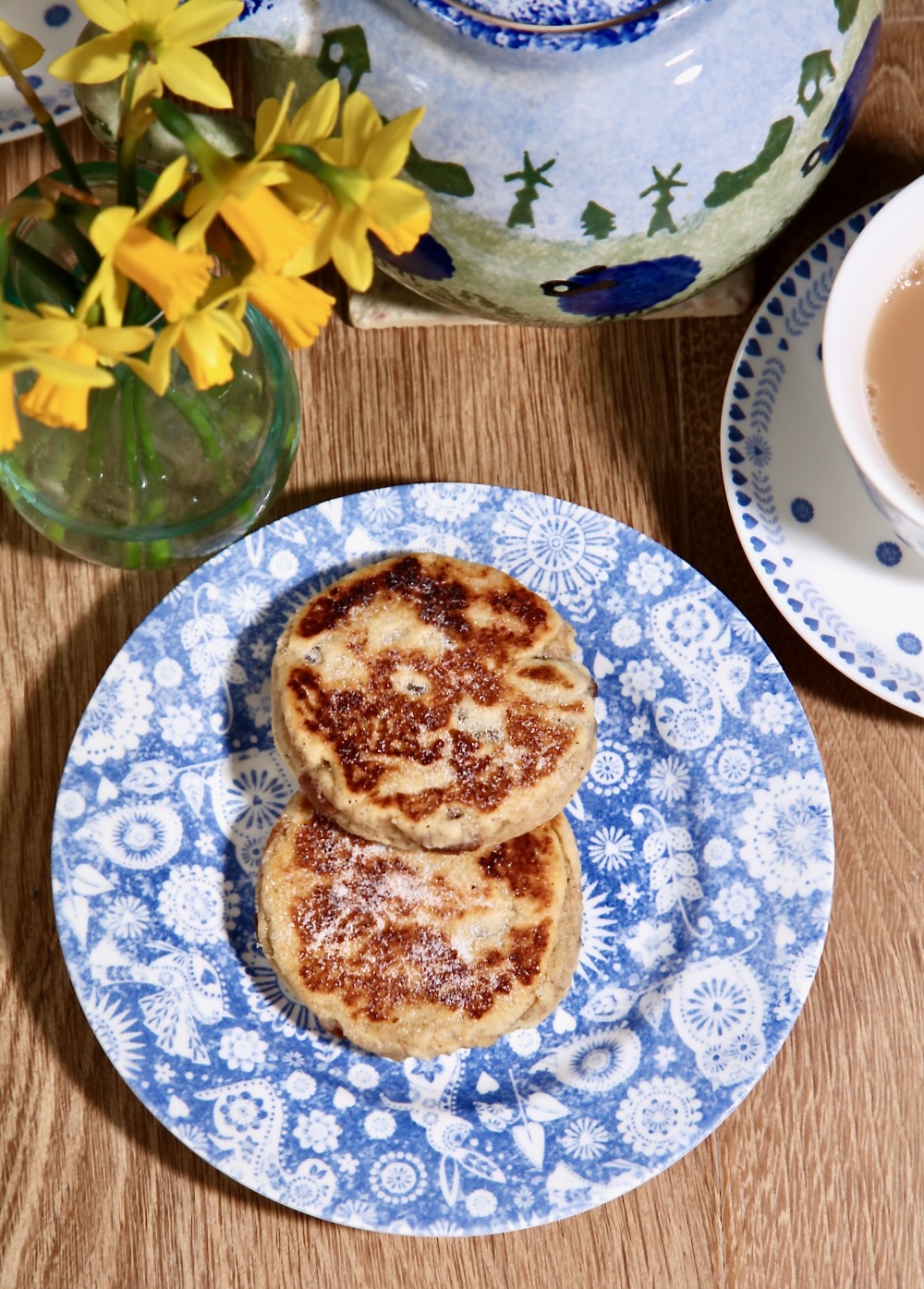 Two Welsh cakes on a blue patterned plate. A cup of tea, a teapot and a vase of daffodils are in the background.