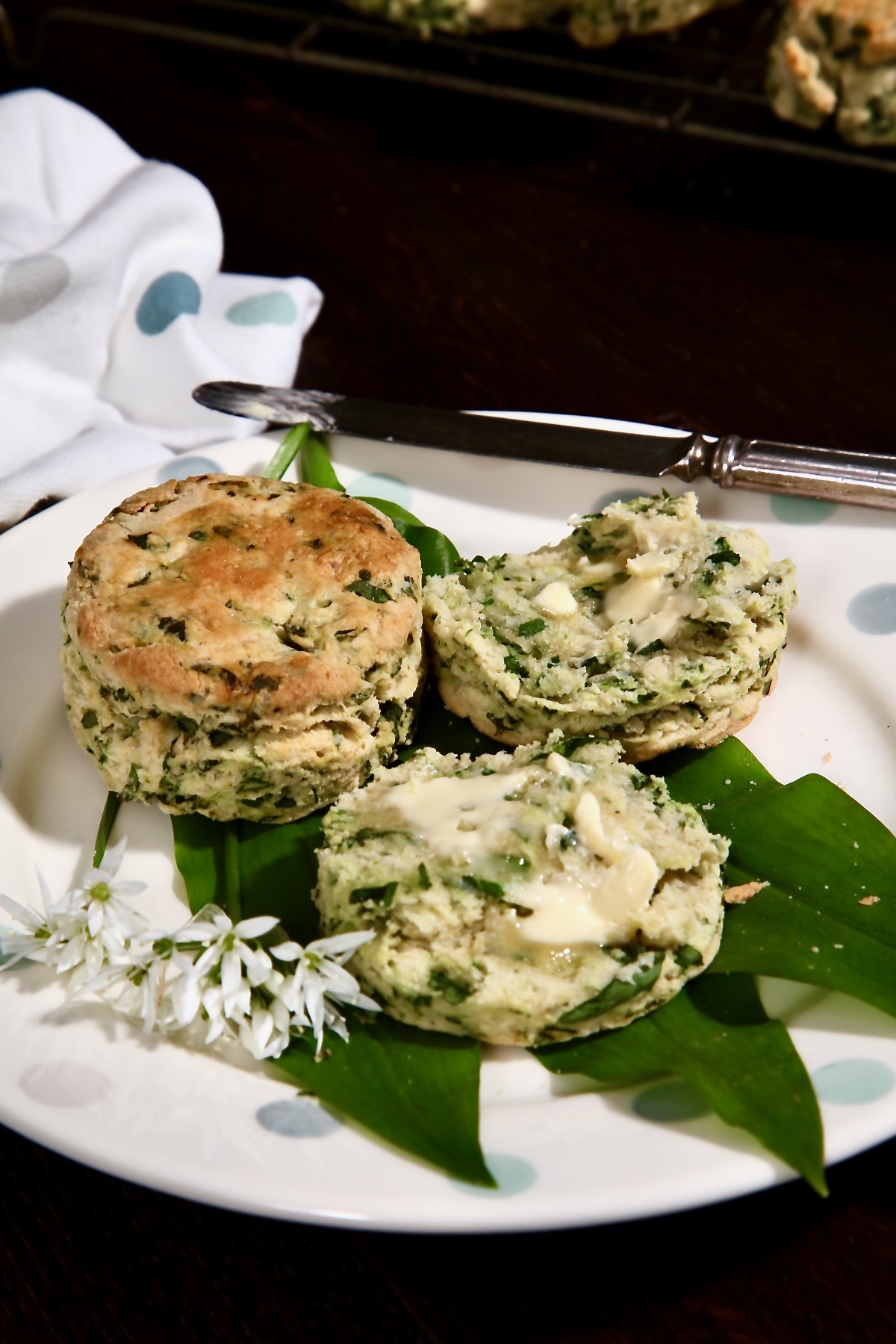 Fresh wild garlic scones are shown on a spotty plate. They are lying on garlic leaves and flowers. One is cut open and spread with melting butter.