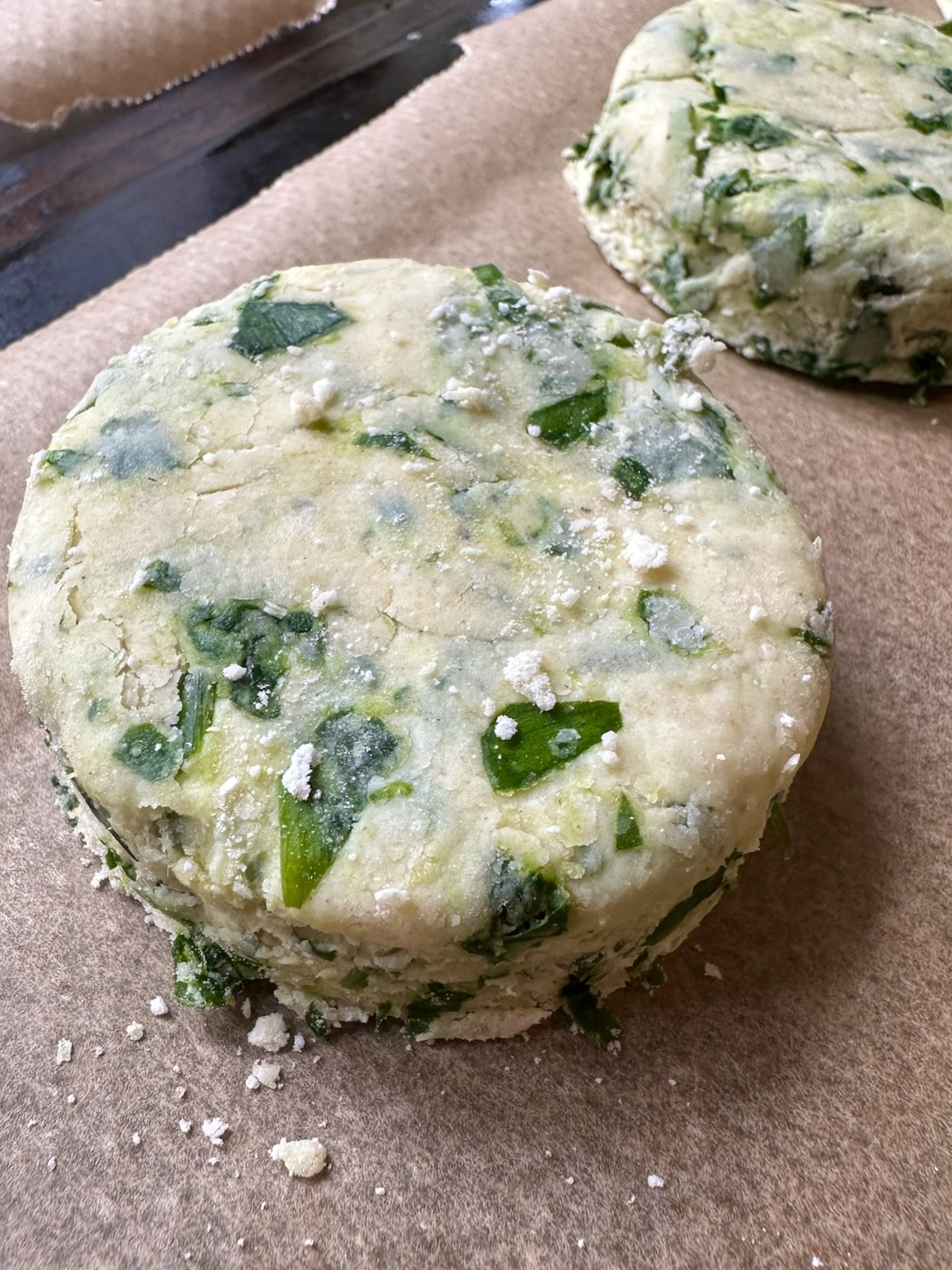 An uncooked wild garlic scone is shown on a baking parchment lined tray, ready for the oven. It is flecked with fresh green wild garlic.