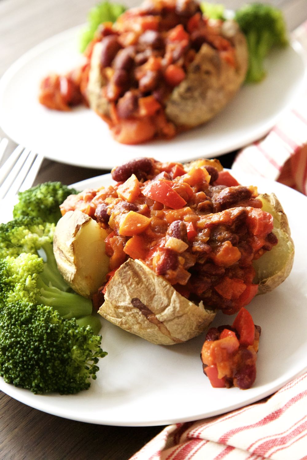 A baked potato is split open and filled with vegan vegetable chilli. It is on a white plate with steamed broccoli. There is another plate of the same, blurred, in the background
