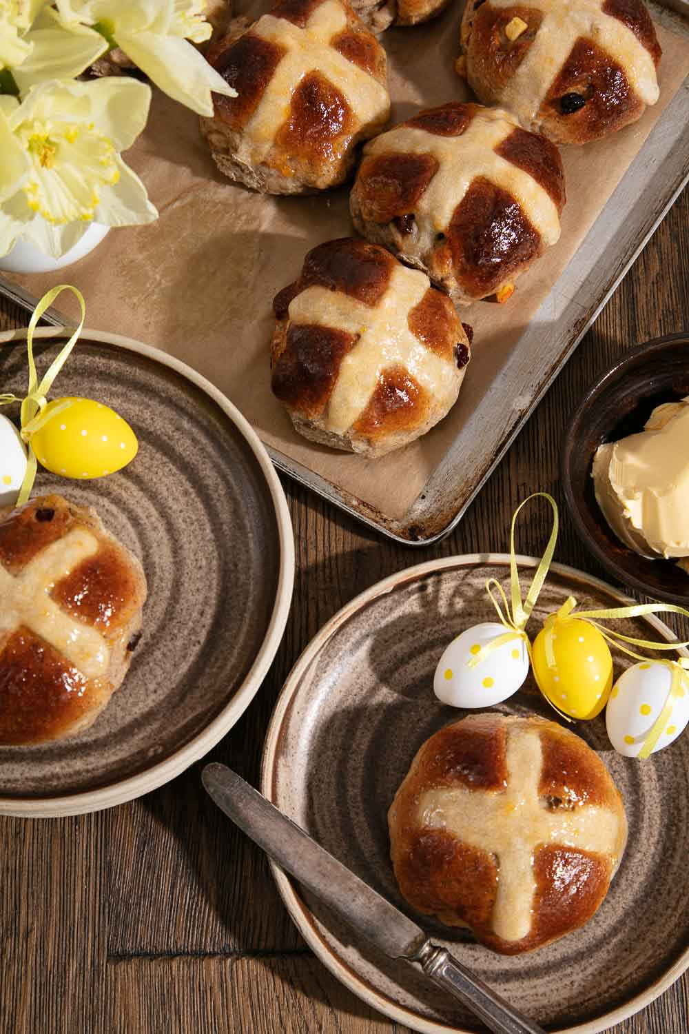 Homebaked Vegan Hot Cross Buns on a baking tray. Two buns on plates with Easter egg decorations. 