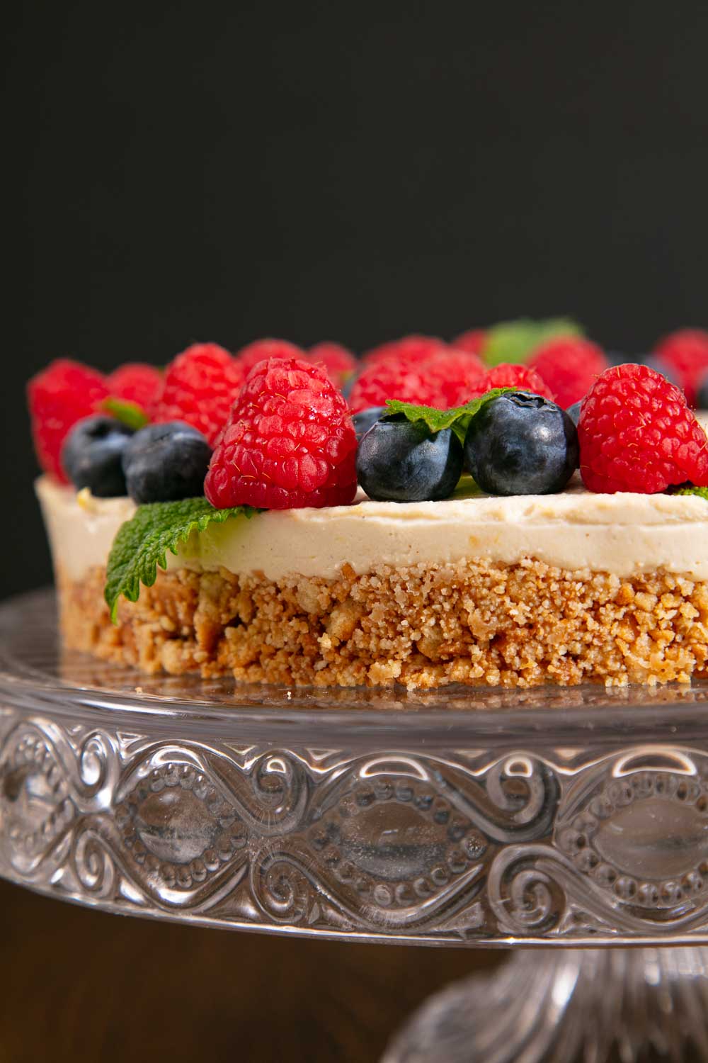 vegan Lemon tart topped with raspberries, blueberries and lemon balm, sitting on a glass cake stand. 