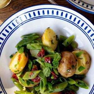 A summer salad of new potatoes, green beans, sun dried tomatoes and fresh herbs presented in a white and blue patterned bowl, a silver spoon and jar of vinaigrette in the background.