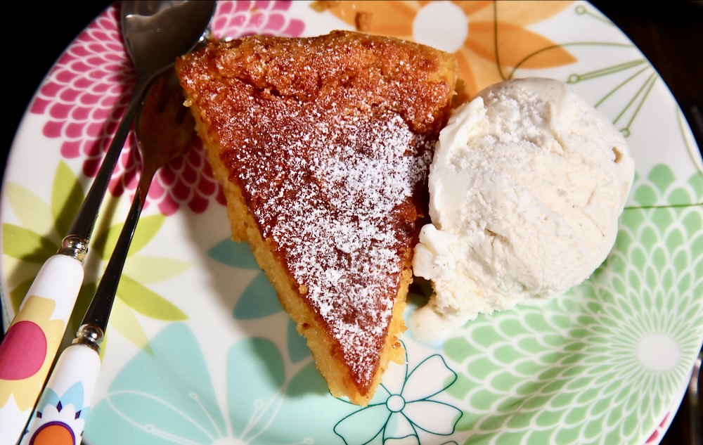 A slice of vegan whole orange cake cake sits on a flowery plate with a scoop of ice cream and a cake fork and spoon.