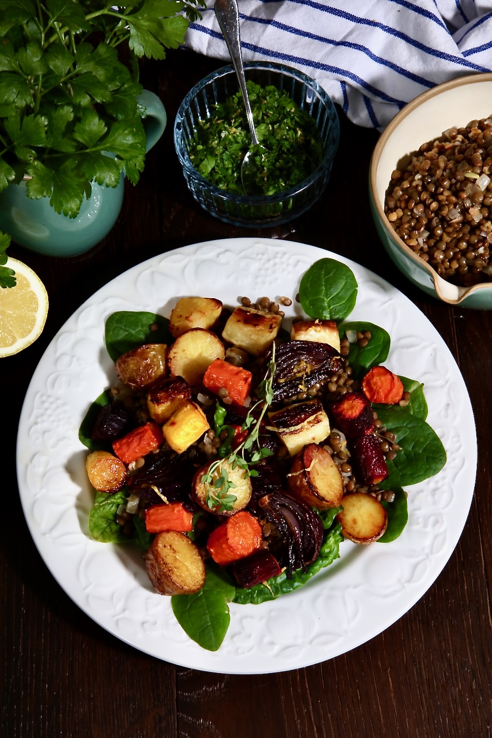 A plate of citrus roasted root vegetables lying on a bed of baby spinach leaves is pictured. Behind it can be seen half a lemon, a small green jug of flat leaved parsley, a glass dish of gremolata and a green vintage pouring jug of cooked green lentils.
