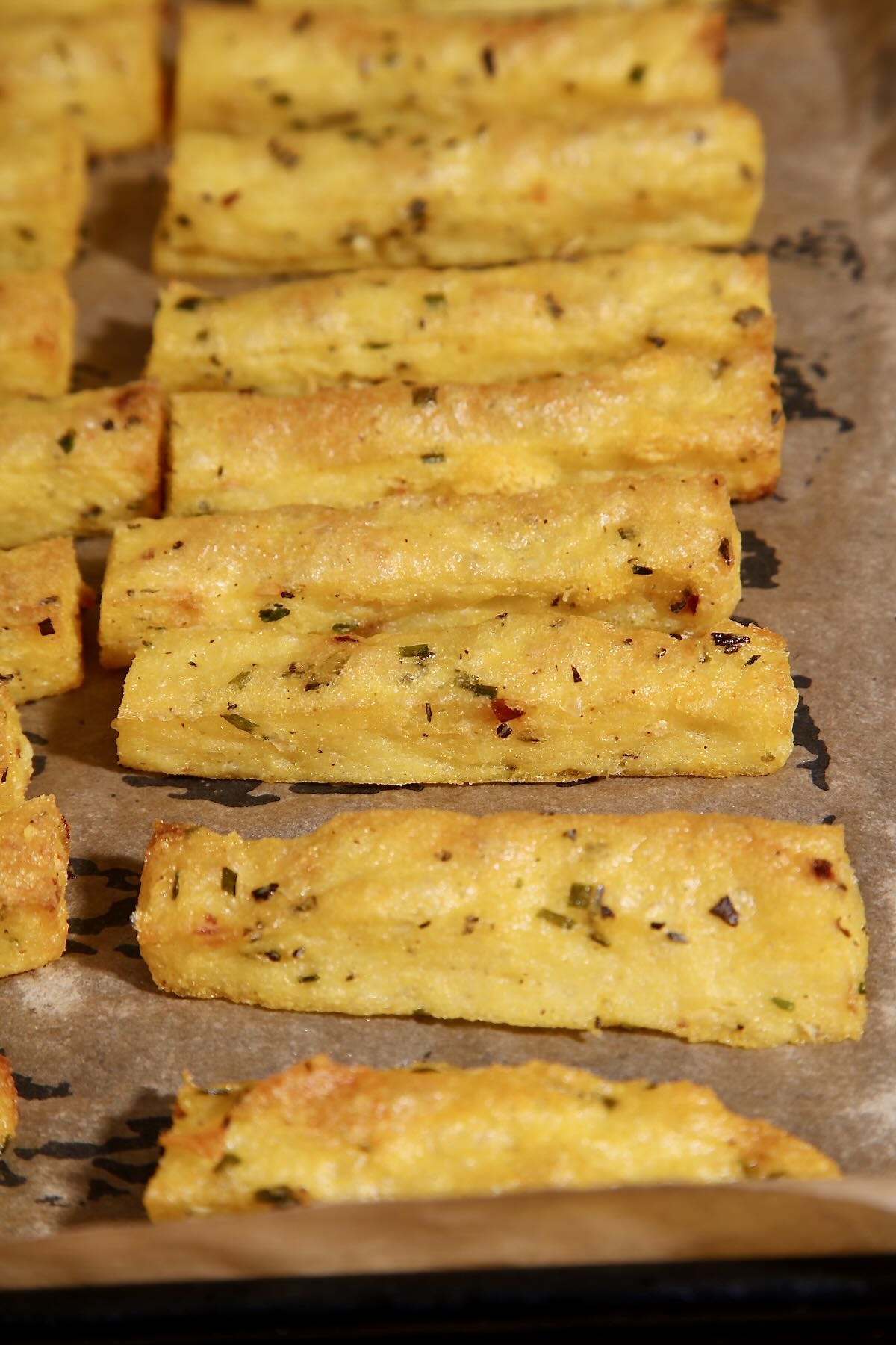 A tray of baked polenta fries. The fries are flecked with rosemary and chilli, they are crisp and golden brown. They are sitting on a parchment lined baking tray.