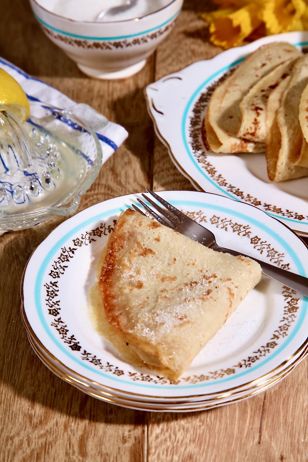 A folded pancake is shown on a vintage plate. The plate is white with a blue stripe and a gold edge. In the background is another plate of the same design with more folded pancakes on it. A glass lemon squeezer full of juice can also be seen. 