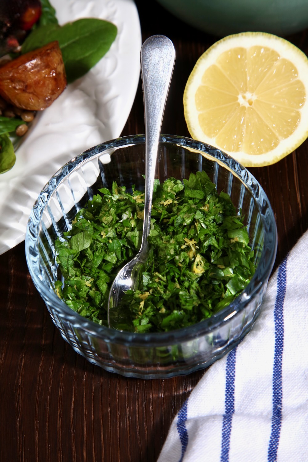 A glass bowl of gremolata with a glimpse of a plate of roasted vegetables in the background along with half a lemon. A blue and white striped cloth can just be seen in the foreground. 