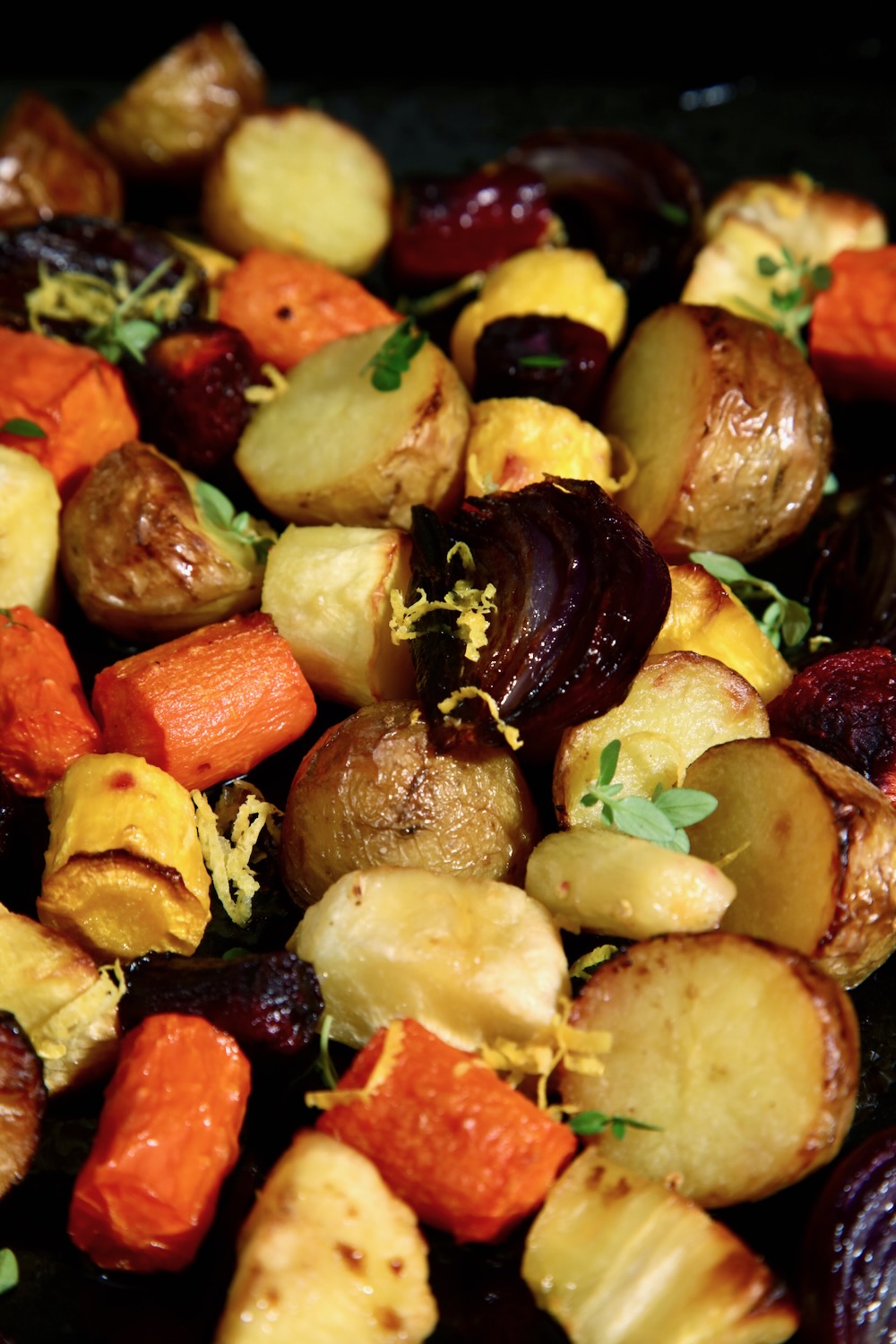 A tray of roasted root vegetables is pictured. Carrots, parsnips and potatoes are cut into bite sized pieces and slices of red onion are scattered throughout. There is a scattering of lemon zest and lemon thyme leaves as a garnish.
