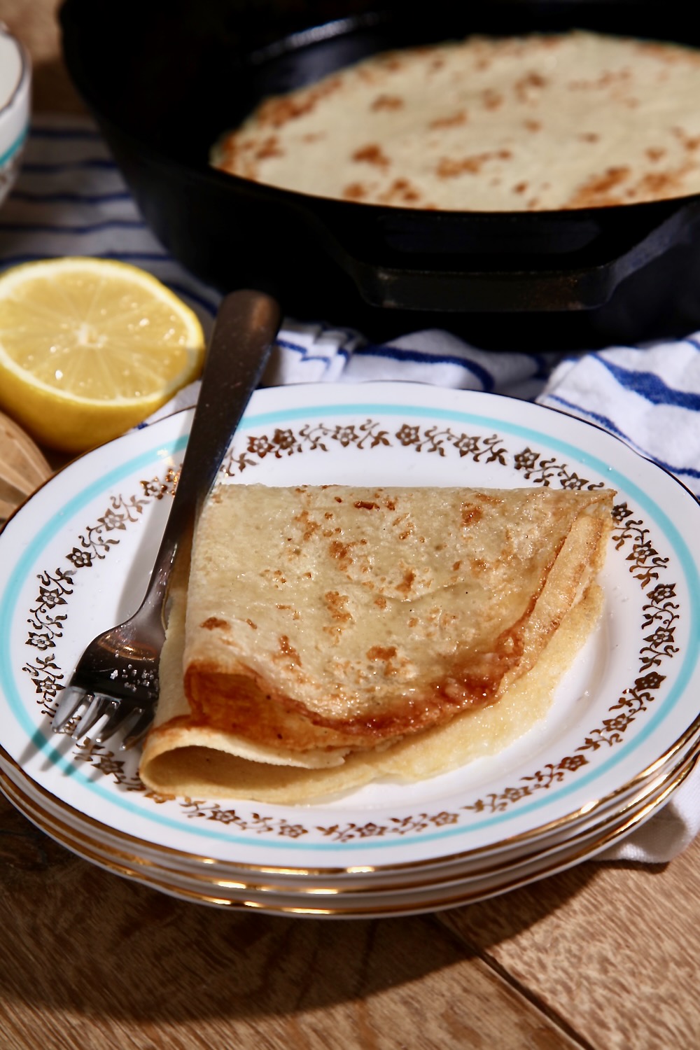 A thin vegan pancake (also known as crêpes_ is shown, folded, on a vintage tea plate. The plate has a turquoise line running around the edge and gold leaf prints within that circle. There is a stainless steel fork next to the pancake. In the background can be seen half a lemon, a cast-iron pan with another pancake inside it and a blue and white striped tea towel.