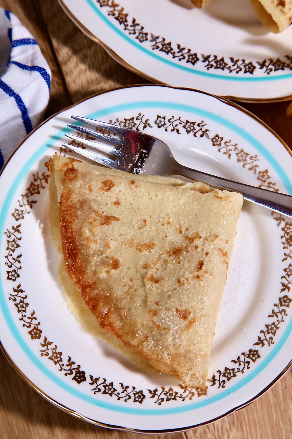 A thin vegan pancake (also known as crêpes_ is shown, folded, on a vintage tea plate. The plate has a turquoise line running around the edge and gold leaf prints within that circle. There is a stainless steel fork on the plate next to the pancake and another plate with pancakes on it can just be glimpsed in the background. 