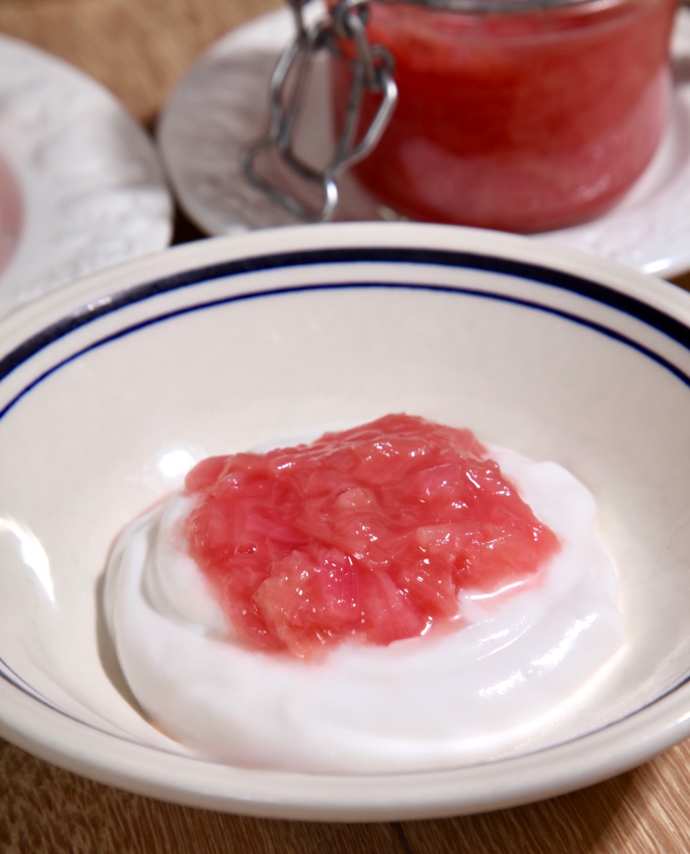 A spoonful of pink rhubarb compote is shown sitting on a generous amount of plain yoghurt in a white bowl with a blue stripe.
