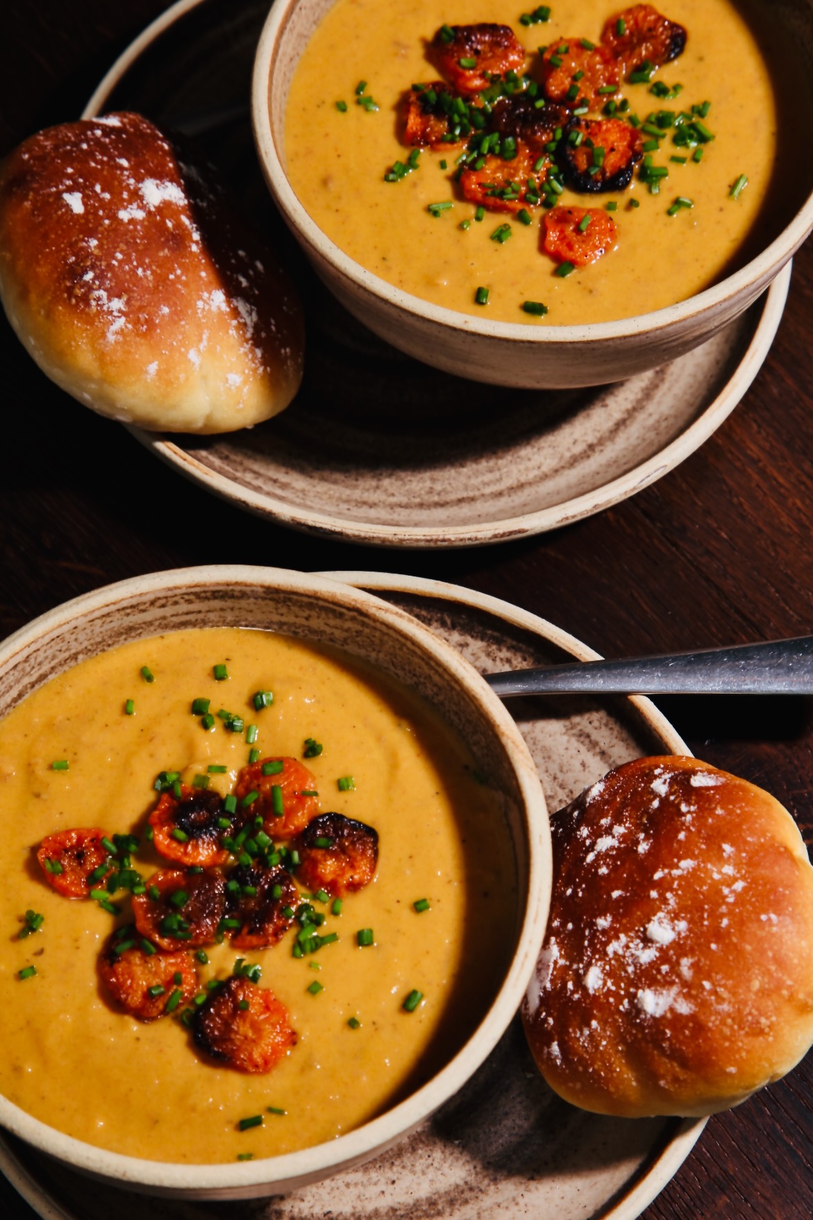 Two rustic bowls full of spiced roasted carrot and borlotti bean soup. They are topped with slices of roasted carrot and chopped chives. They sit on matching plates and are accompanied by fresh, floury bread rolls.