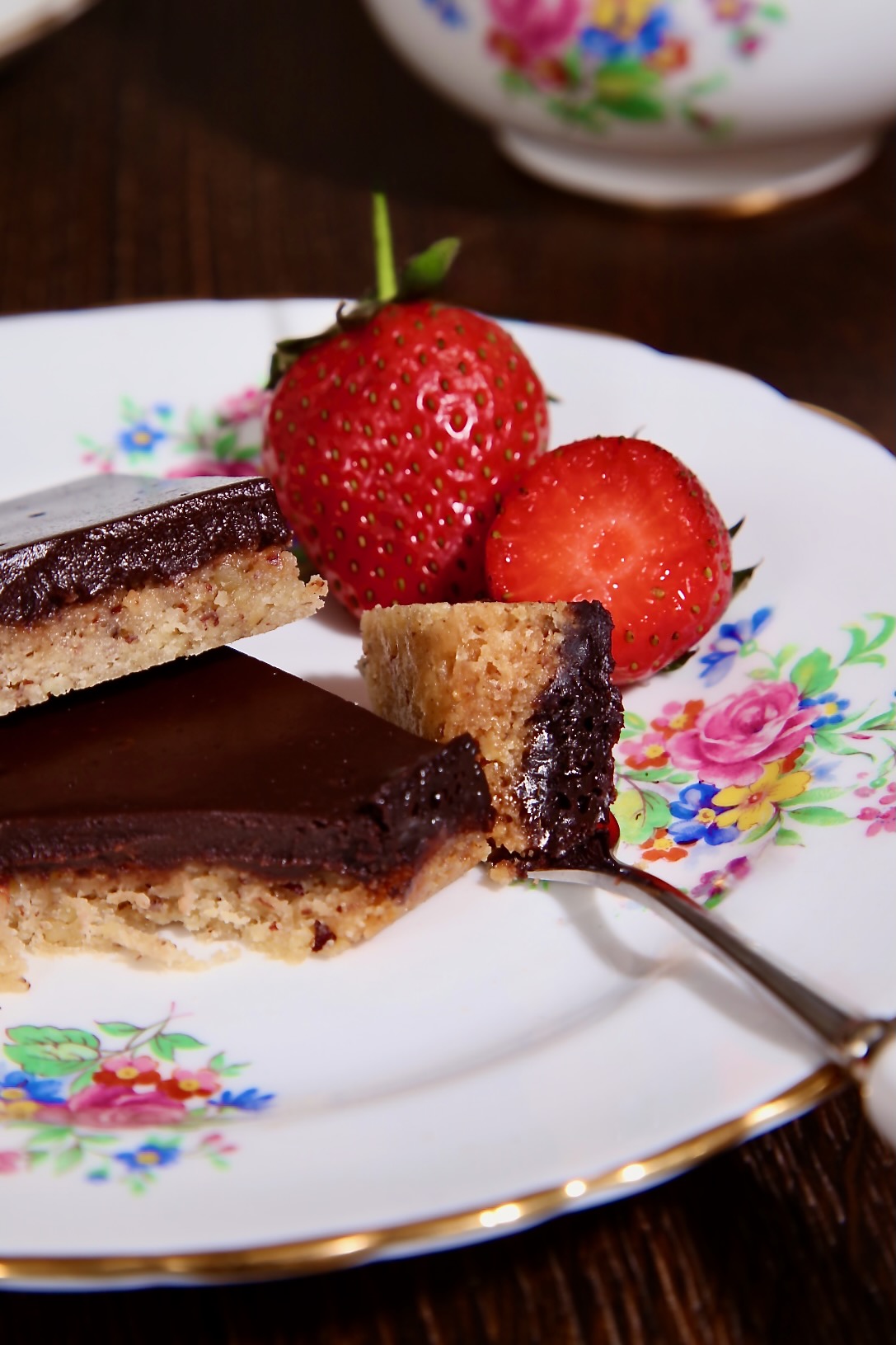 Two chocolate pecan bars sit on a floral patterned plate. A piece of the dessert is sitting on a cake fork, ready to eat. There are two juicy strawberries in the background. 